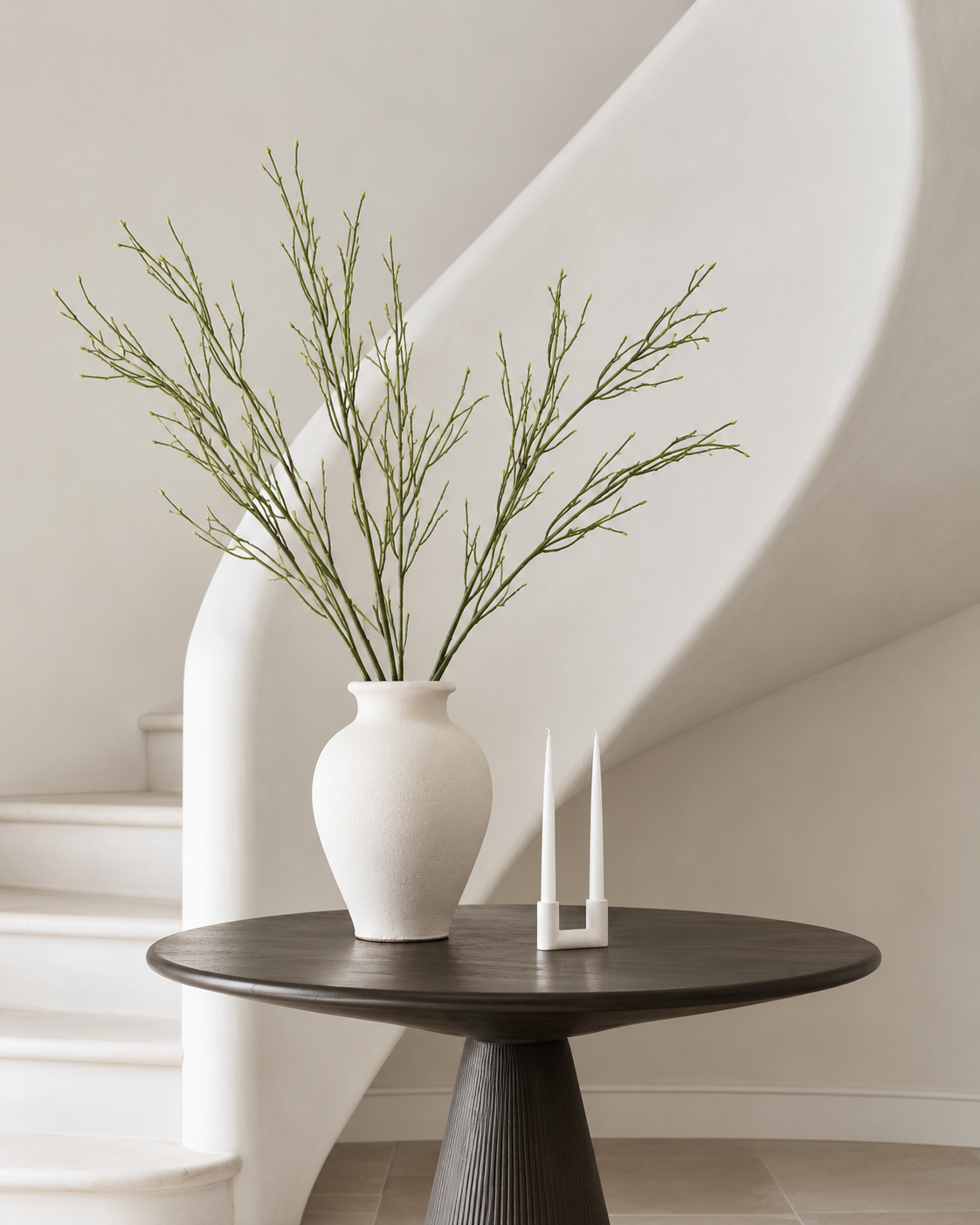 Saltmarsh faux stems in a white textured vase on a round dark timber table beside a curved white staircase