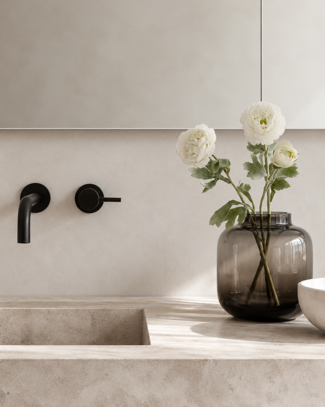 White faux ranunculus stems in a smoky glass vase on a stone bathroom vanity beside black tapware