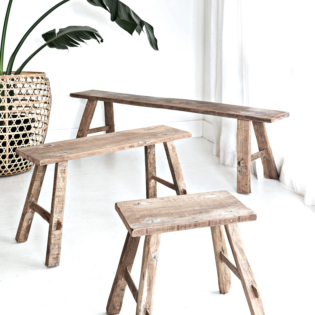 Three teak benches in graduated lengths grouped beside a woven basket and plant