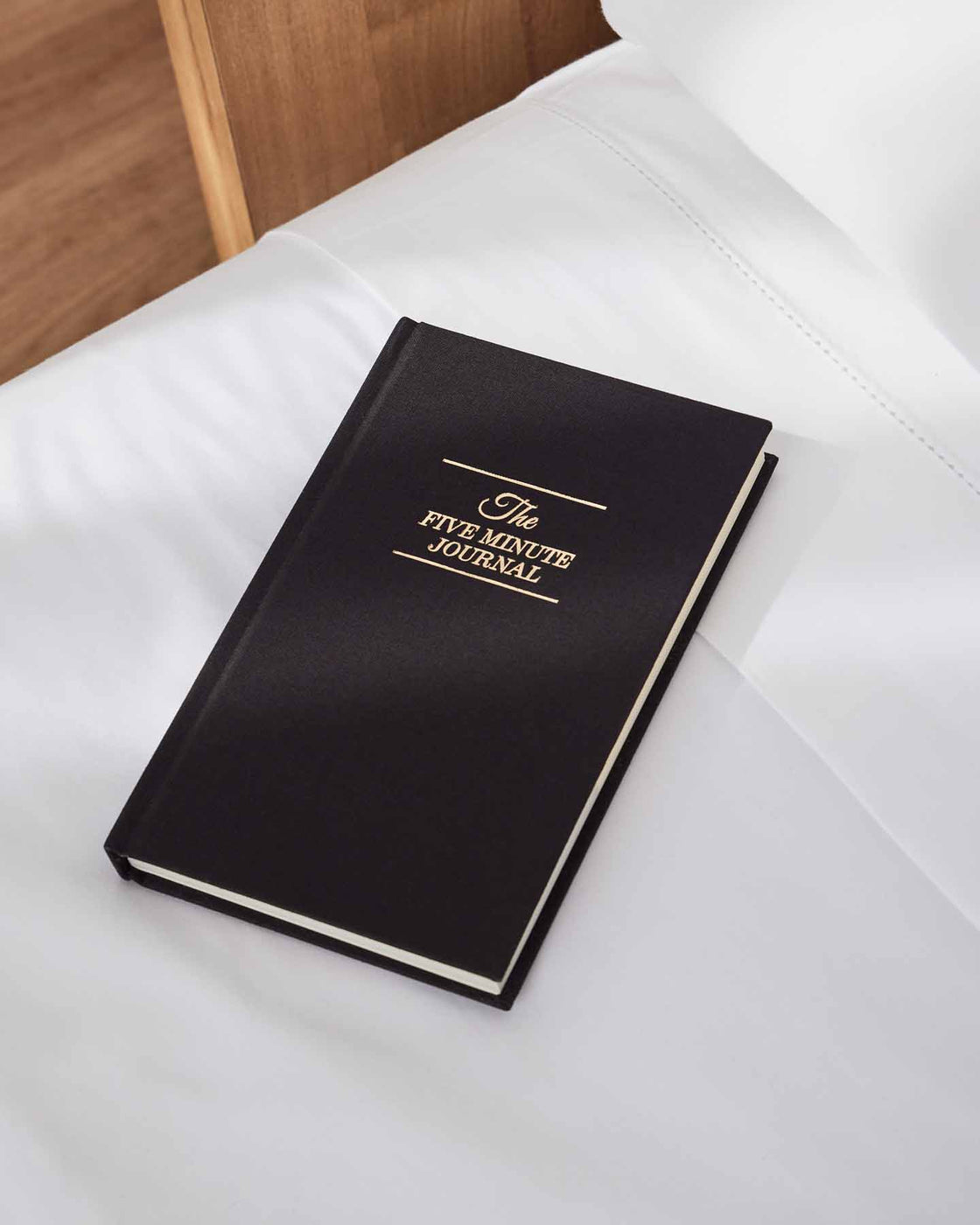 Black Five Minute Journal on white bedding beside a timber bedhead