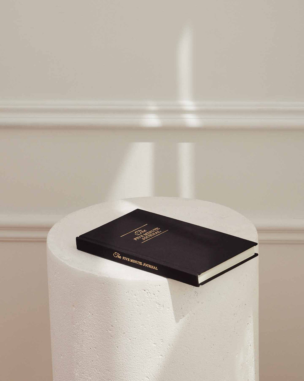 Black Five Minute Journal on a white plinth in soft shadow