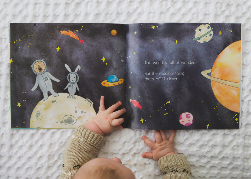 A baby is holding a Olive + Page's "IN A WORLD FULL OF MAGIC" book with a space theme.