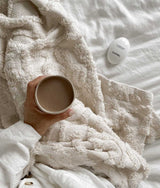 A person holding a cup of coffee on a bed, surrounded by Bengali Collections' Ivory Tufted Throw for ultimate comfort.