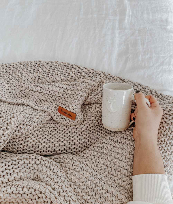 A person holding a cup of coffee on a bed with a Bengali Collections CHUNKY KNIT OATMEAL THROW blanket.