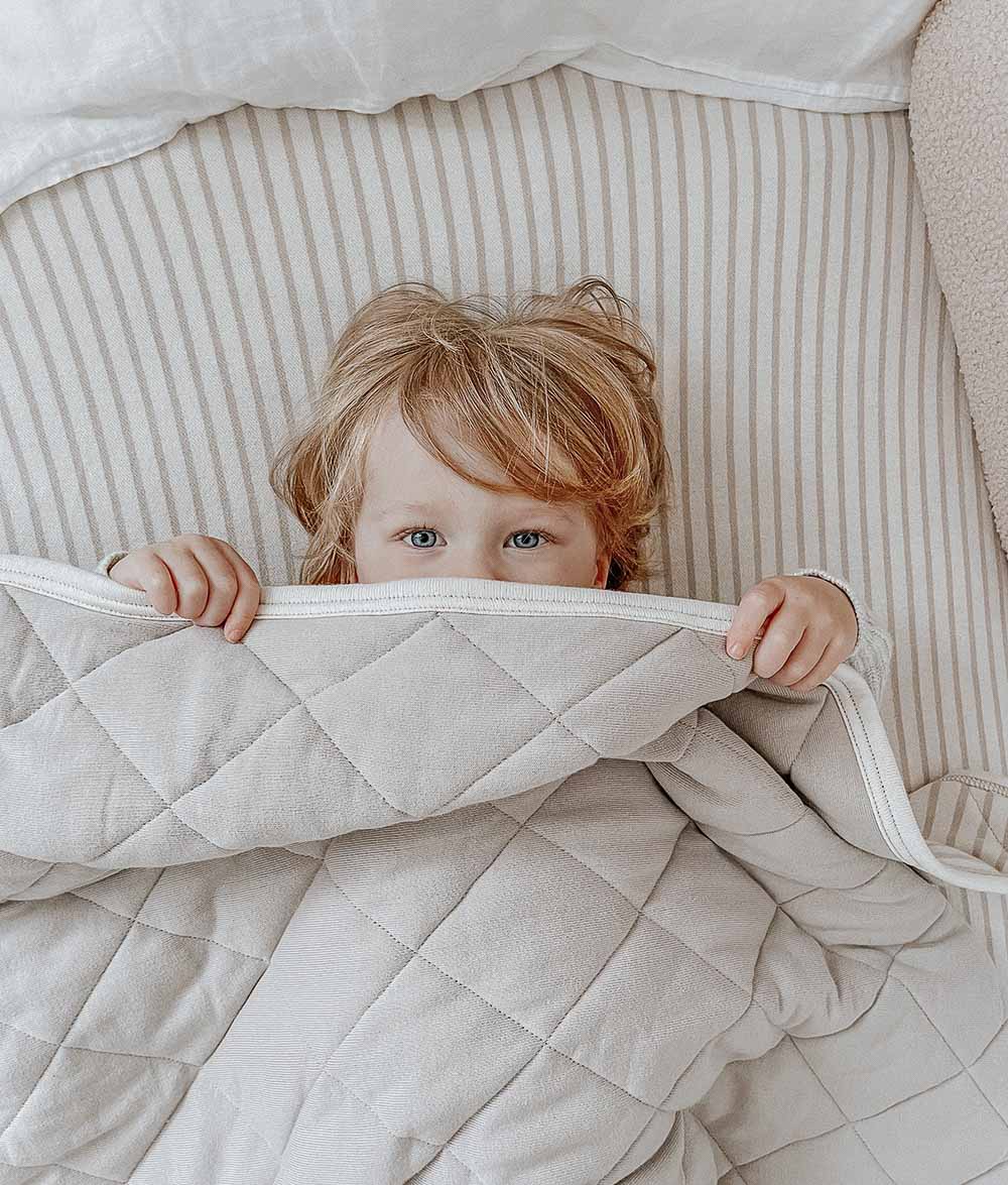 Child peeking over a quilt while lying on a greige pinstripe jersey cotton fitted sheet