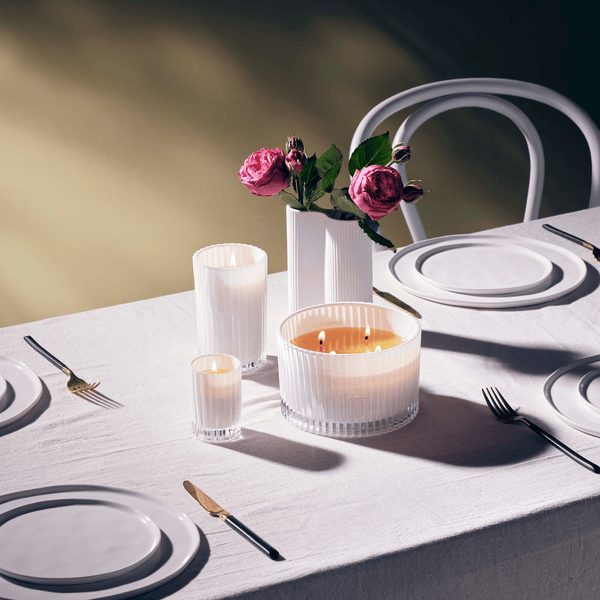 A white table setting adorned with a beautiful arrangement of candles and flowers, including an Ecoya Celebration | White Musk & Warm Vanilla Grand Candle.