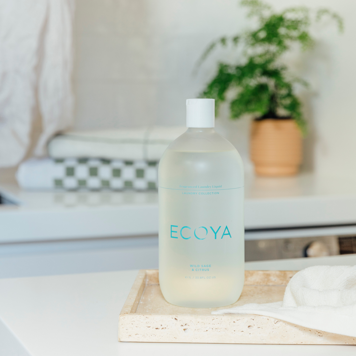 Ecoya laundry liquid on a wooden tray with folded linen and a potted plant on a white bench