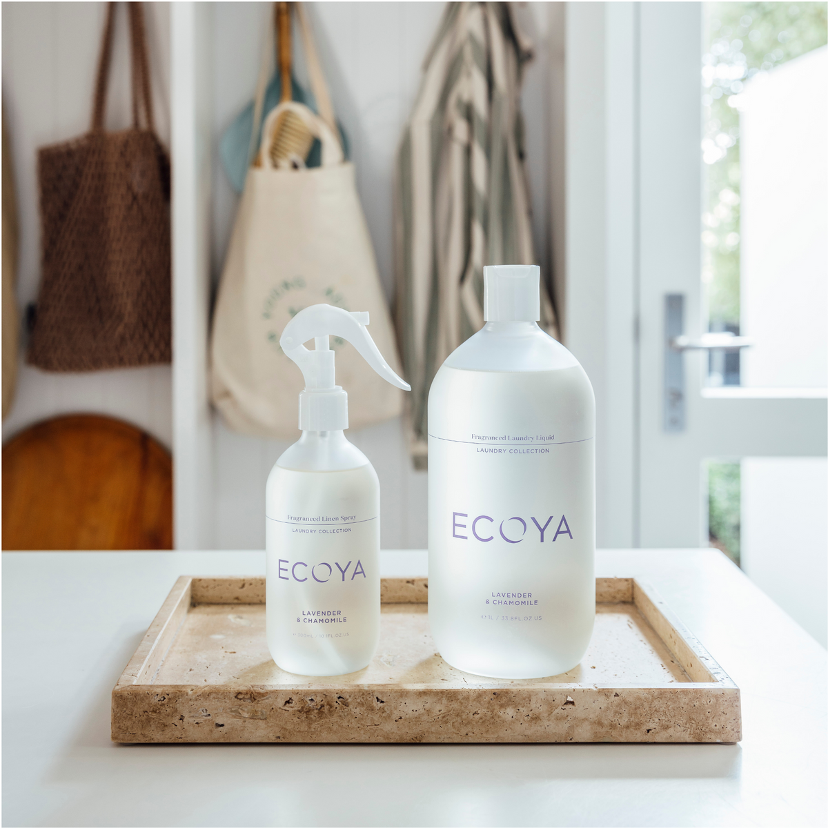 Ecoya linen spray and laundry liquid on a wooden tray by a doorway with bags and towels hanging