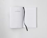 An open I Am The Hero Of My Own Life notebook on a white surface with a black ribbon, perfect for envisioning your ideal life.