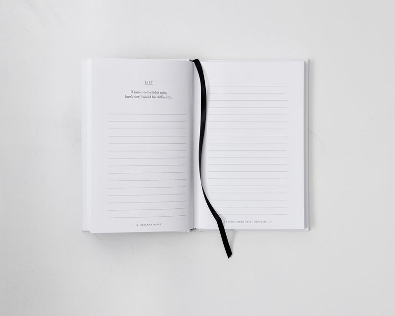 An open I Am The Hero Of My Own Life notebook on a white surface with a black ribbon, perfect for envisioning your ideal life.