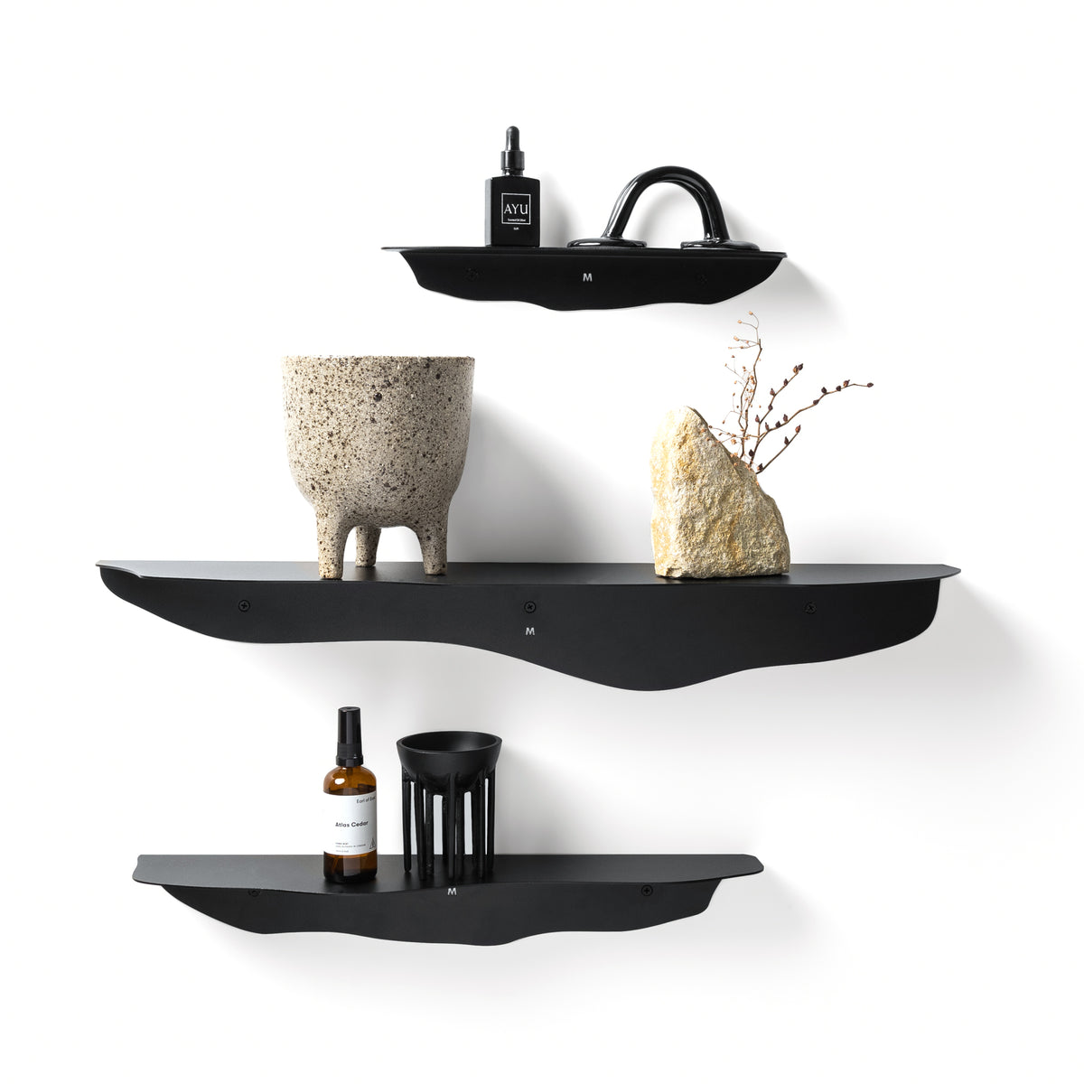 Three black wavy shelves staggered on a white wall with a stone planter, vase and candle holder