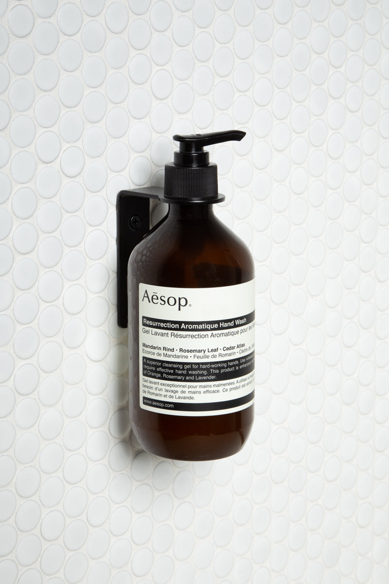 A bottle of aesop soap hanging on a bathroom wall with the Made of Tomorrow FOLD Bottle Holder ∙ Black.