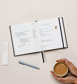 A hand holding the List Maker | Long Sticky Note Set | Pack of Two notebook from Emma Kate Co brand and a cup of coffee, perfect for planner enthusiasts and list makers. The notebook is filled with neatly organized pages and colorful sticky notes to help you stay organized and on track.