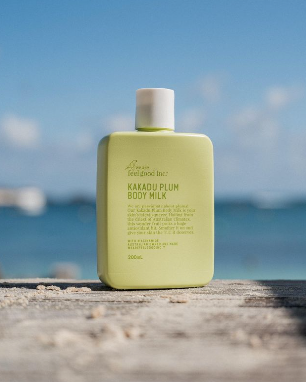 A bottle of We Are Feel Good Inc. Kakadu Plum Body Milk sitting on a wooden table in front of the ocean.