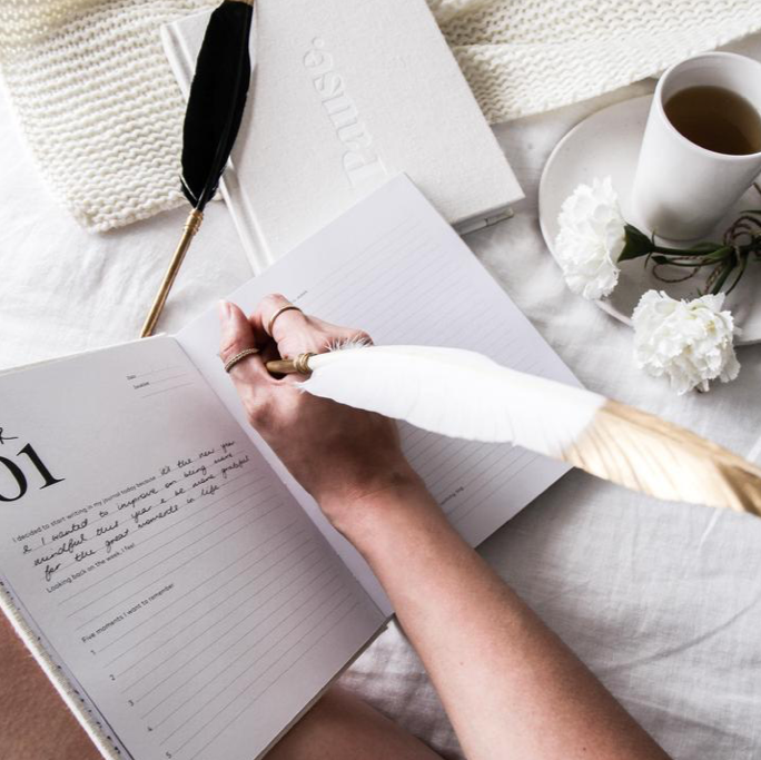 Hand writing in a journal with the white Coco feather pen beside coffee and white flowers