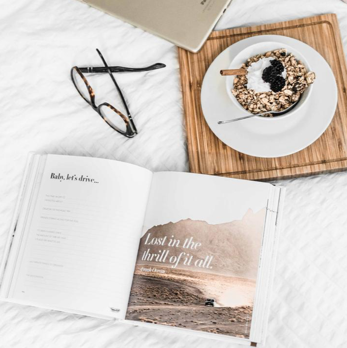 An open book with coffee, glasses and a laptop on a bed, showcasing the stylish Life's A ROADTRIP - Luxe Edition by AXEL & ASH range.