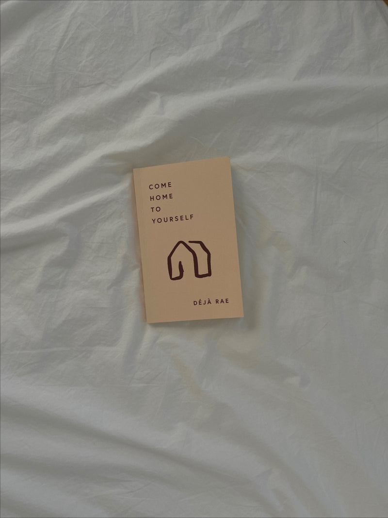 A "Come Home to Yourself | Déjà Rae" design book resting on a plush bed, adorned with an enchanting illustration of a cozy house, perfectly capturing the essence of solitude and relationships. This beautifully curated product is brought to you by Thought Catalog.