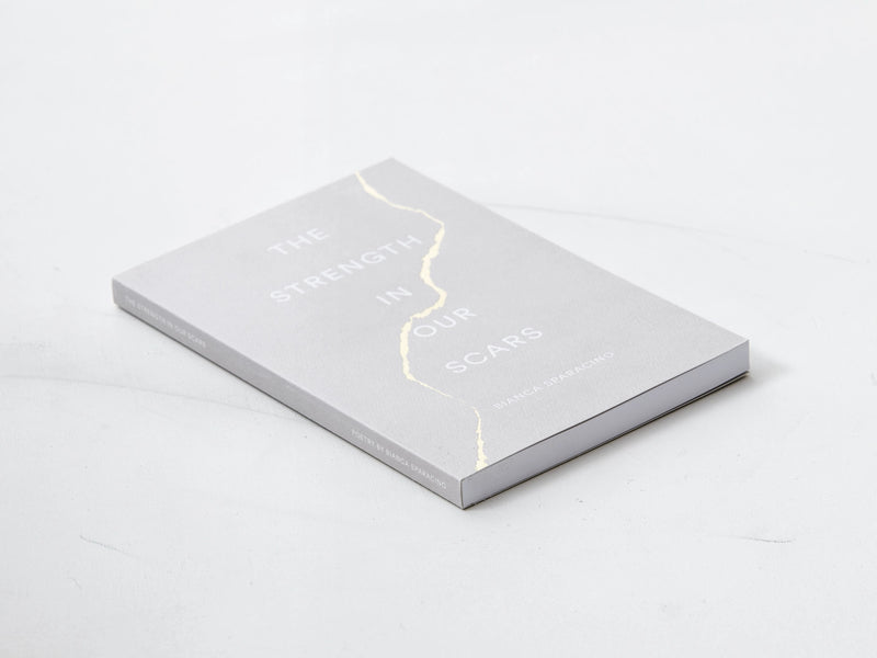 A "The Strength In Our Scars | Bianca Sparacino" book with a gold foil cover on a white surface.