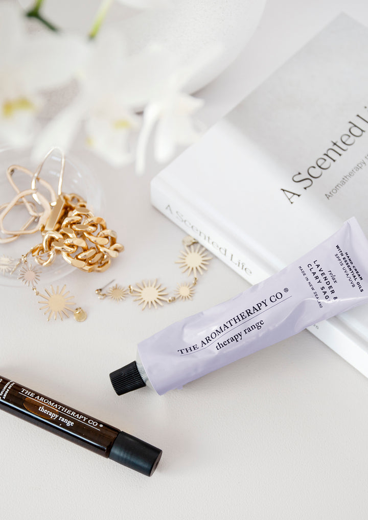 Lilac hand cream tube and an amber rollerball beside gold jewellery and a book with white orchids