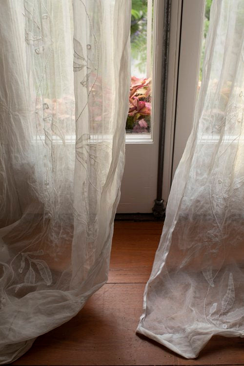 White sheer curtains in a Victoria Alexander home, gracefully casting shadows on a wooden floor in front of a window.