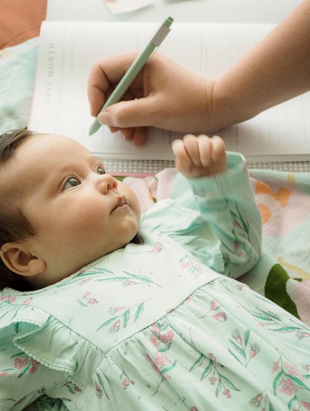 A baby lying on a bed while someone writes in the Write To Me Baby Journal
