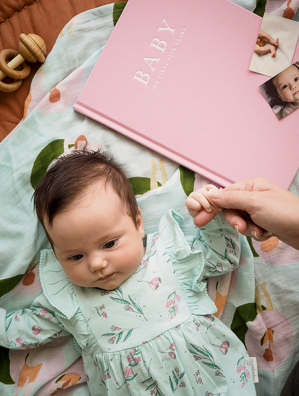 Baby lying on a floral blanket beside the pink Write To Me Baby journal and photos