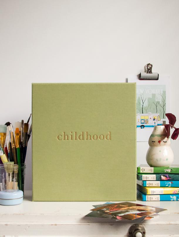 Sage linen Childhood journal on a kids' desk with paintbrushes, stacked books and loose photos
