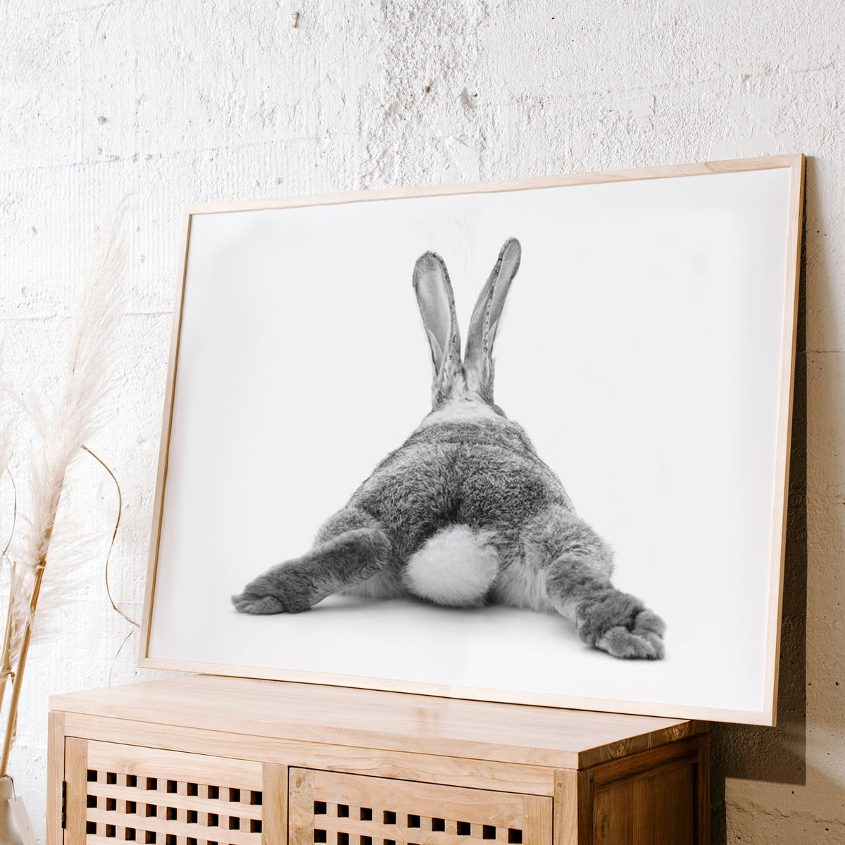 Bunny Bum print in a white frame leaning on a rattan cabinet against a textured wall