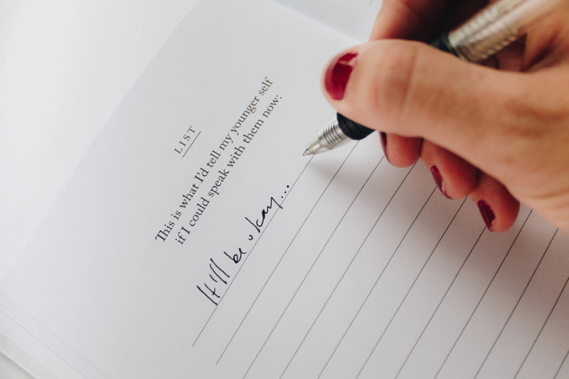 A woman writing in a notebook with a pen, envisioning her ideal life using Thought Catalog's product, "I Am The Hero Of My Own Life" by Brianna Wiest.