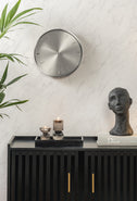Silver Jewel clock above a black sideboard with a sculptural bust and palm leaves