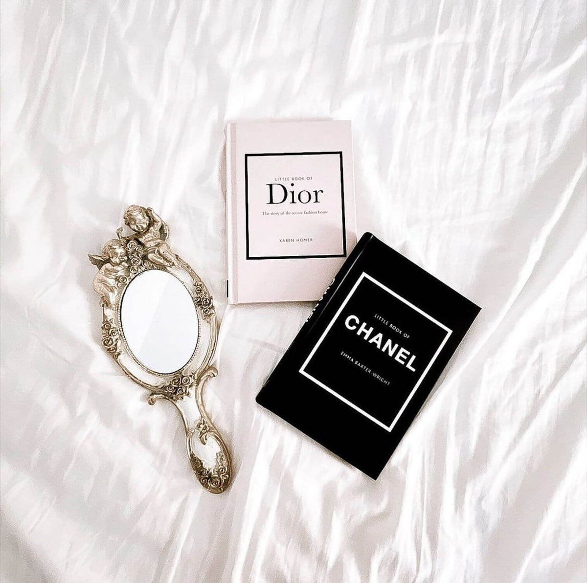 Little Book of Chanel and Little Book of Dior with a vintage hand mirror on white bedding