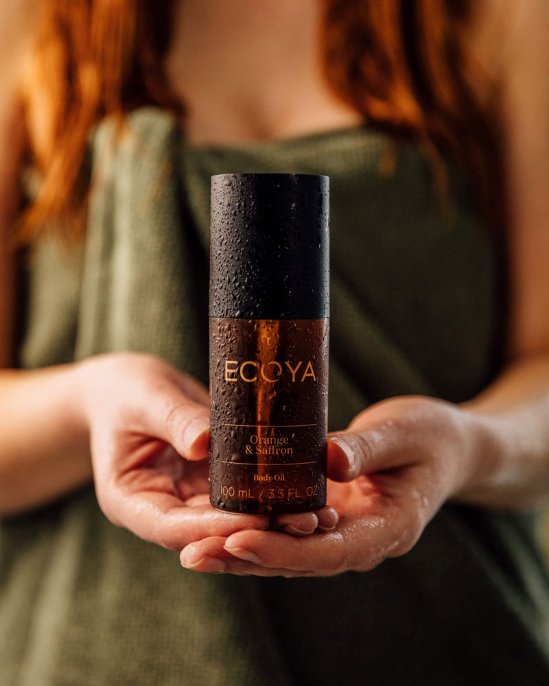 A scandinavian-inspired woman holding a bottle of Ecoya Limited Edition | Orange & Saffron Body Oil, perfect for home design enthusiasts seeking a sophisticated fragrance.