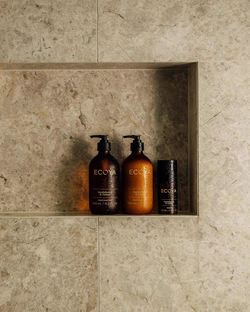 Two bottles of Ecoya Limited Edition Sandalwood & Amber Hand & Body Wash, providing home fragrance, sit on a shelf in a bathroom.