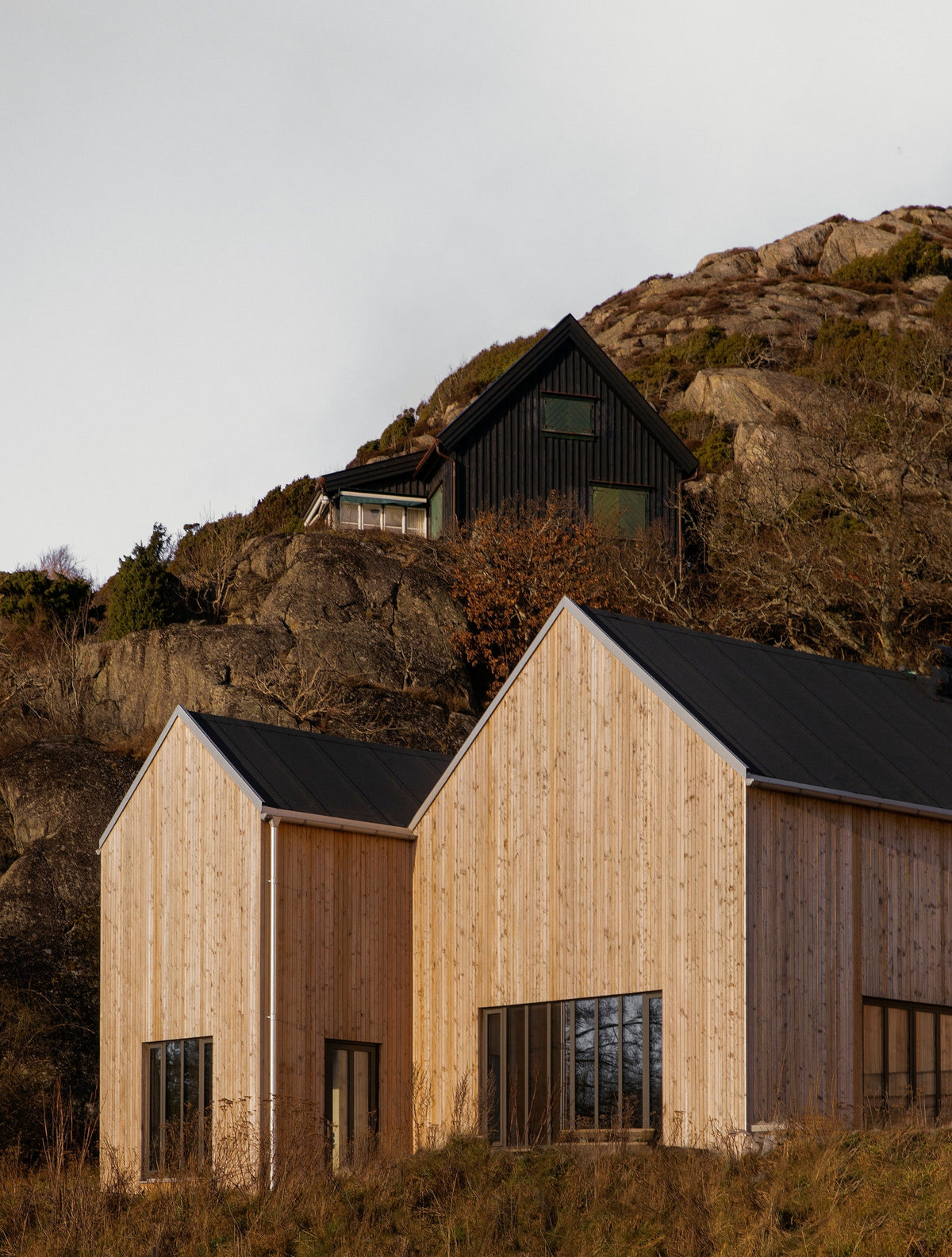 Timber clad gabled cabins on a rocky coastal hillside below a dark house