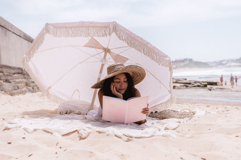 A woman on a wellness journey reading the Healthy Nourished Soul book under an umbrella on the beach published by Epicurean Publishing.