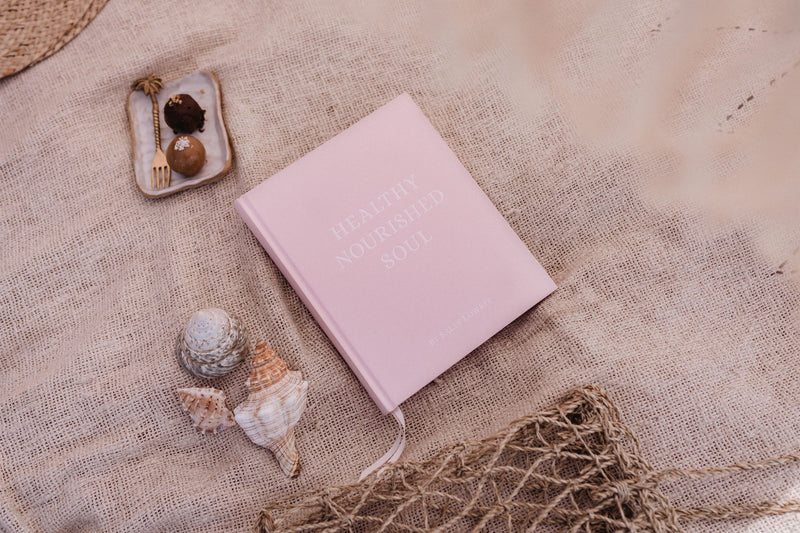 A Healthy Nourished Soul - Book by Epicurean Publishing on a beach blanket with seashells and a bag.