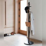 An Umbra Pillar Coat Rack in a hallway with a coat hanging on it for storage.