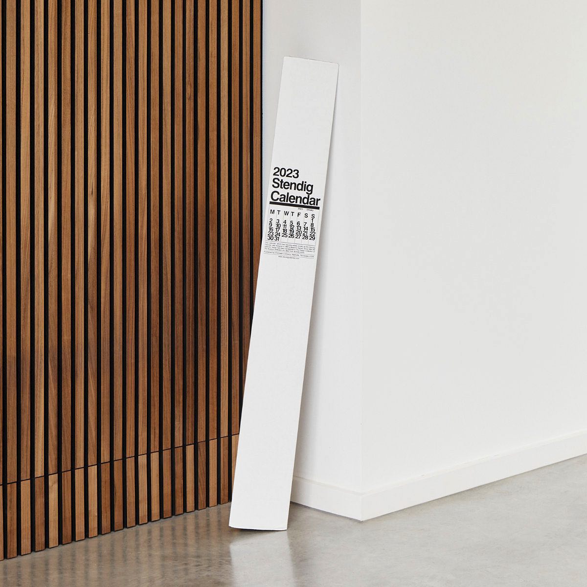 The Stendig Calendar in its branded packaging tube leaning against a timber slat wall, showing the rolled oversized format