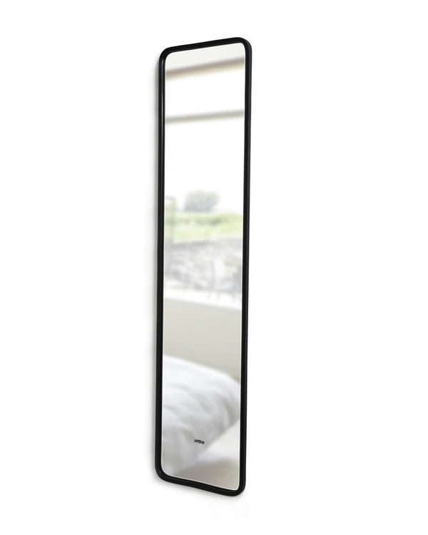 A HUB LEANING MIRROR - BLACK from the Umbra range hanging on a wall next to a bed.