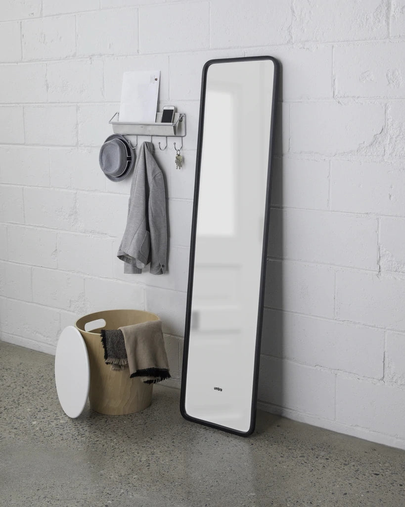 Umbra Hub leaning mirror in black against a white brick wall beside a stool