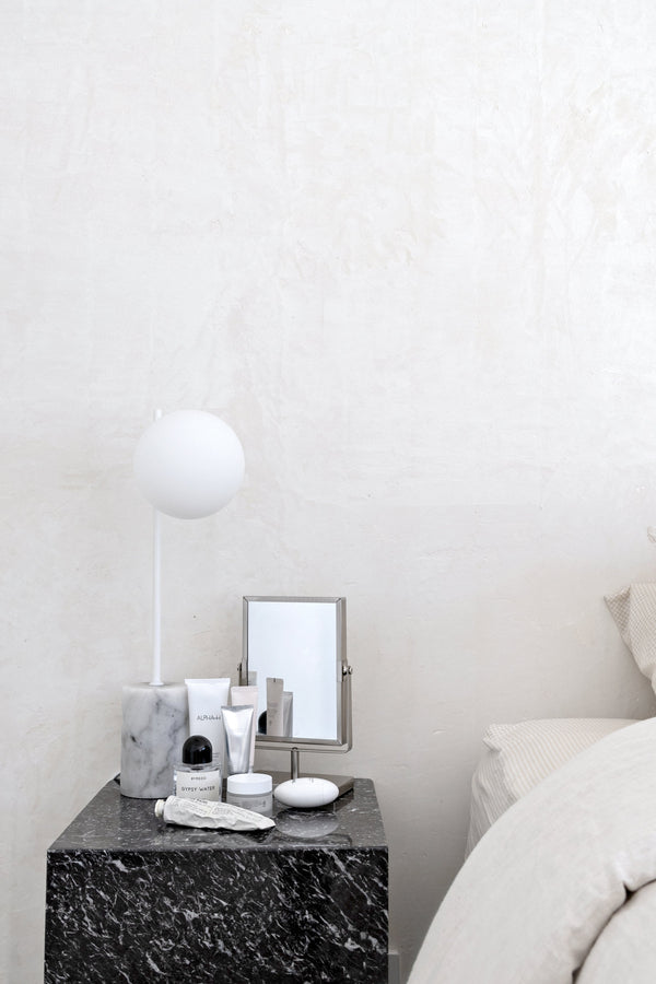 Minimalist bedside scene with a marble lamp, a small mirror, curated skincare products, and a black marble side table, set against a textured neutral wall and soft bedding.