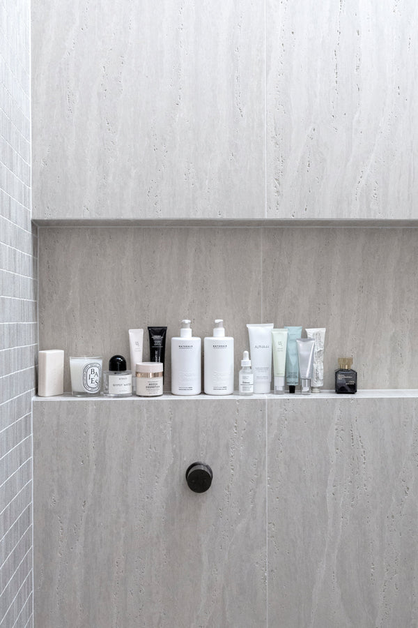 A minimalist shower niche neatly lined with curated skincare and body care products in neutral-toned packaging against soft grey stone tiles.