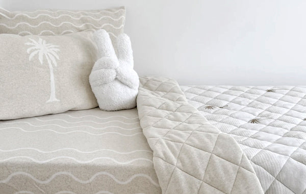 Neutral kids’ bedding with palm tree print, wave pattern sheets, and a plush peace-sign cushion