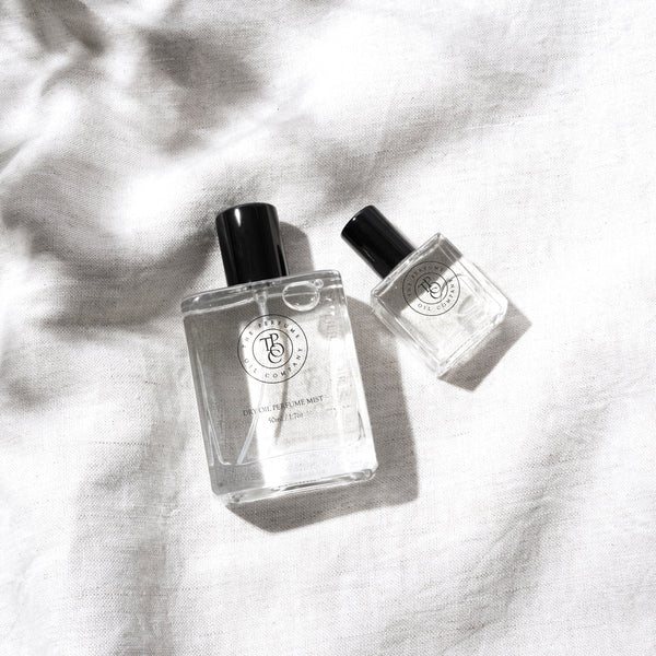Two minimalist glass bottles of The Perfume Oil Company fragrance, including a Dry Oil Perfume Mist and a roll-on perfume, styled on a soft natural linen background with gentle sunlight and shadow detail.