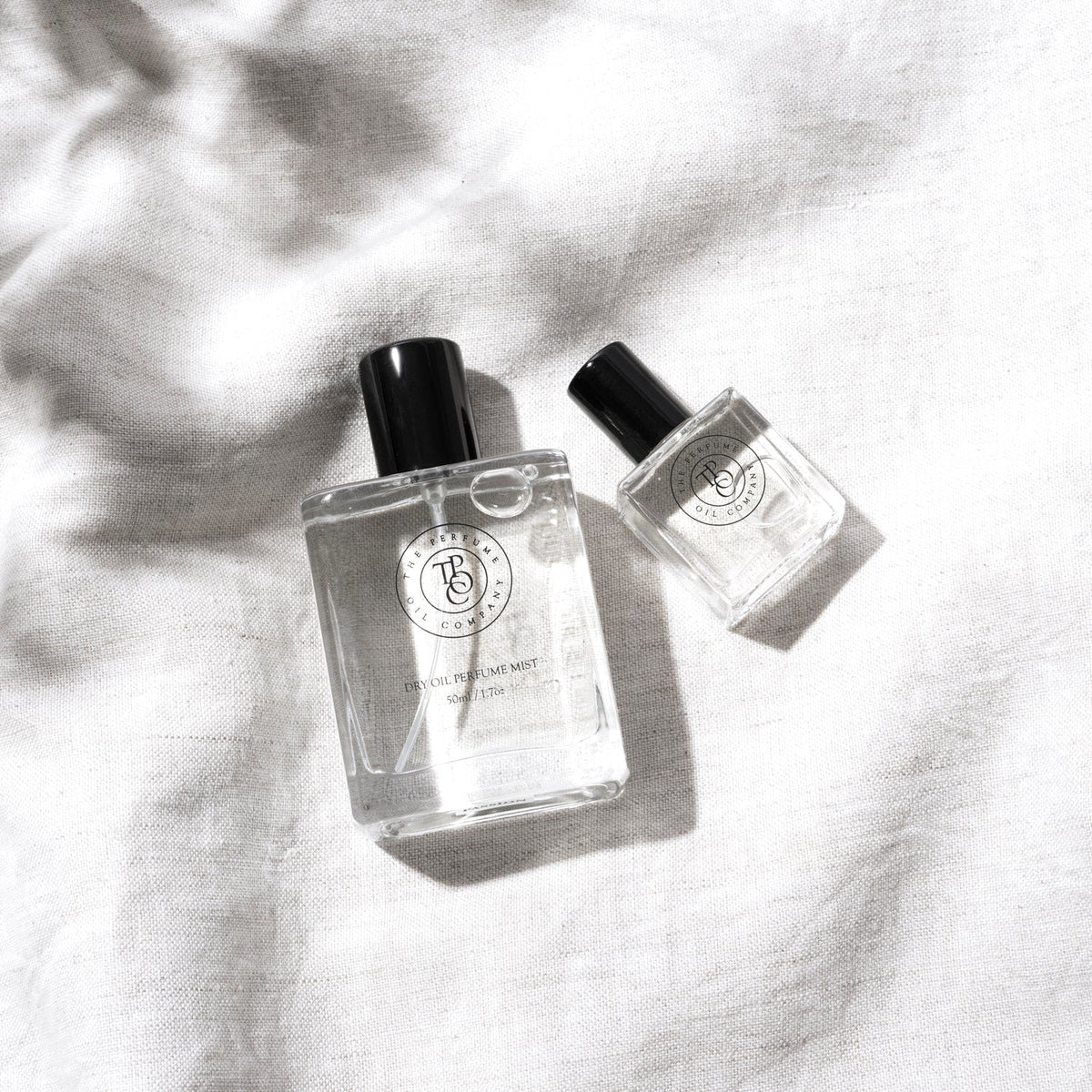 Two minimalist glass bottles of The Perfume Oil Company fragrance, including a Dry Oil Perfume Mist and a roll-on perfume, styled on a soft natural linen background with gentle sunlight and shadow detail.
