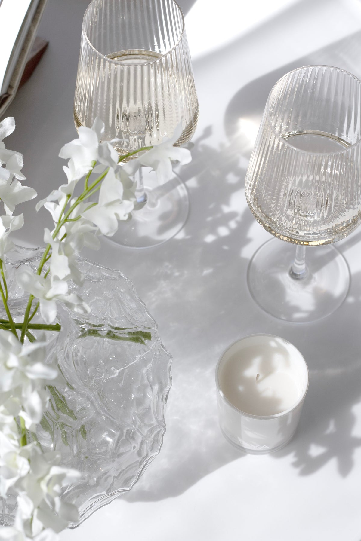 Two fluted wine glasses with white florals, a candle and a glass bowl in soft sunlight