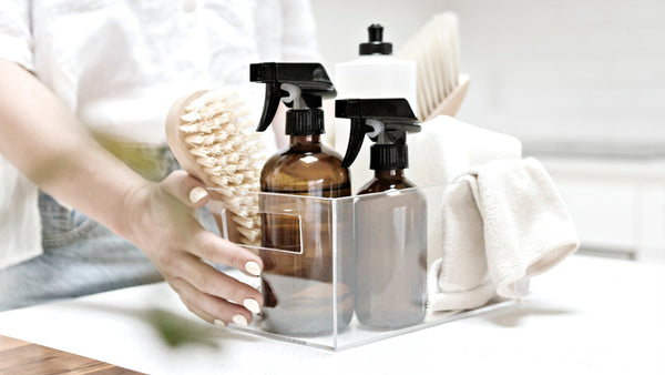 Hand reaching for a minimalist cleaning caddy with amber spray bottles, wooden brush, cloths, and dish soap — a calm, curated toolkit for a fast home reset.