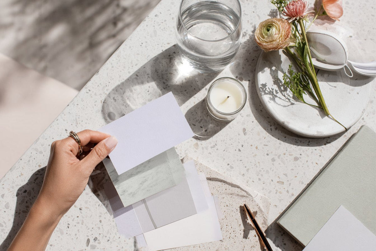 Overhead view of a candle, ranunculus, a glass of water and paper swatches on a terrazzo table
