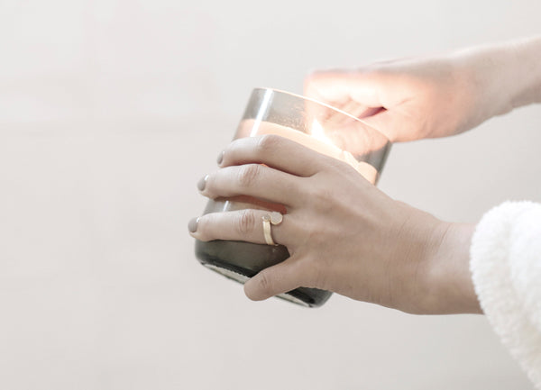 Close-up of hands holding and lighting a softly glowing candle in a neutral-toned glass jar, evoking a cozy and intentional atmosphere for seasonal transitions.