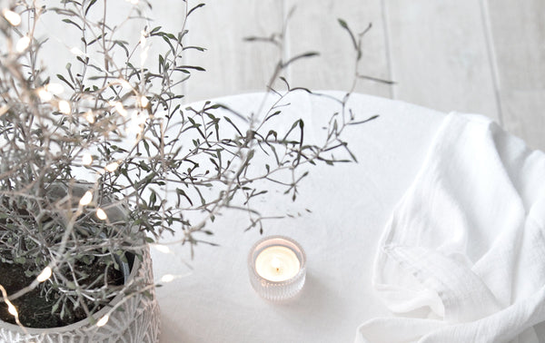 Close-up of a cozy autumn scene featuring a potted plant wrapped in delicate fairy lights, a glowing tealight candle in a ribbed glass holder, and a soft white cloth on a light surface, creating a warm, inviting atmosphere.